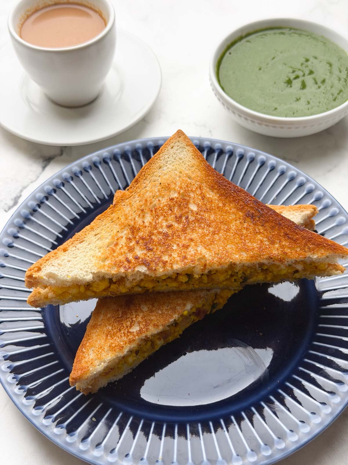 Two paneer bhurji sandwich stacked on a plate with green chutney and paneer bhurji on the side.