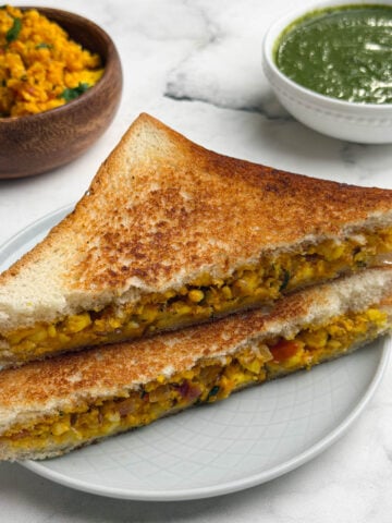 Two paneer bhurji sandwich stacked on a plate with green chutney and paneer bhurji on the side.