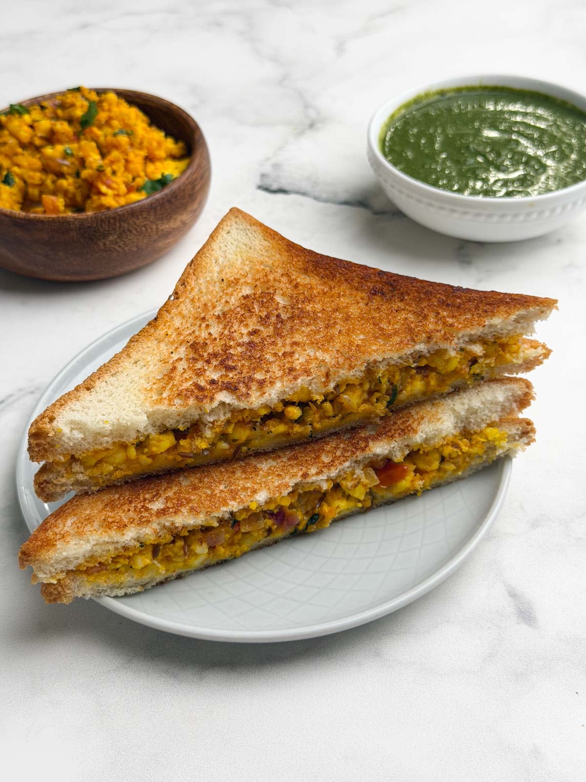 Two paneer bhurji sandwich stacked on a plate with green chutney and paneer bhurji on the side.