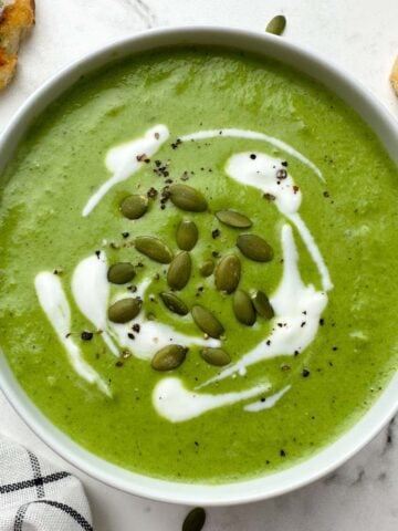 broccoli spinach cheese soup served in a bowl garnished with cream and pumpkin seeds with garlic bread on the side