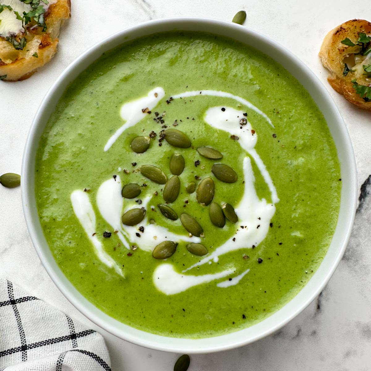 broccoli spinach cheese soup served in a bowl garnished with cream and pumpkin seeds with garlic bread on the side