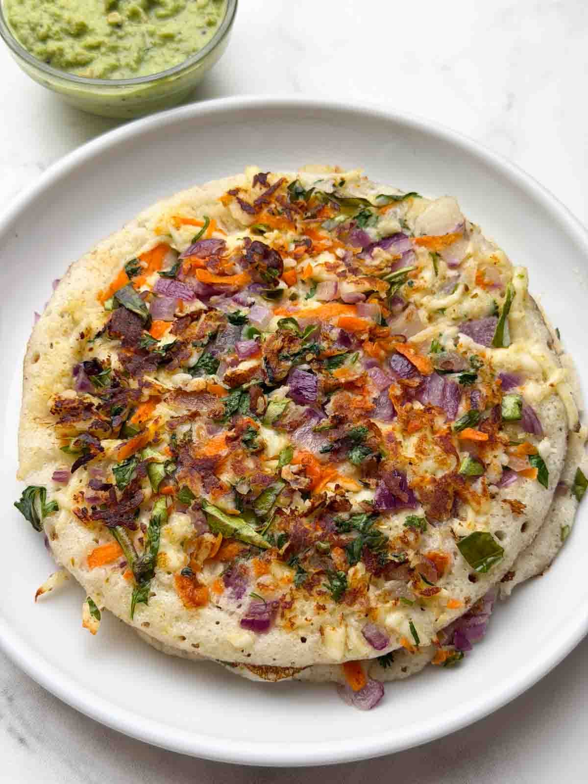 Paneer vegetable uttapams stacked on a white plate with coriander coconut chutney on the side.
