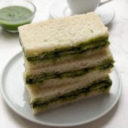 A stack of cucumber chutney sandwiches on a white plate with masala chai and green chutney on the side.
