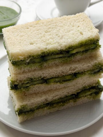 A stack of cucumber chutney sandwiches on a white plate with masala chai and green chutney on the side.