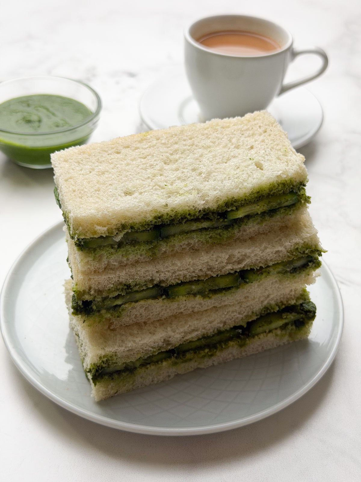 A stack of cucumber chutney sandwiches on a white plate with masala chai and green chutney on the side.