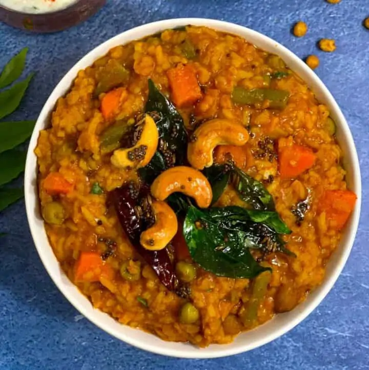 bisi bele bath served in a white bowl topped with tempering and raita on side along with some boondi