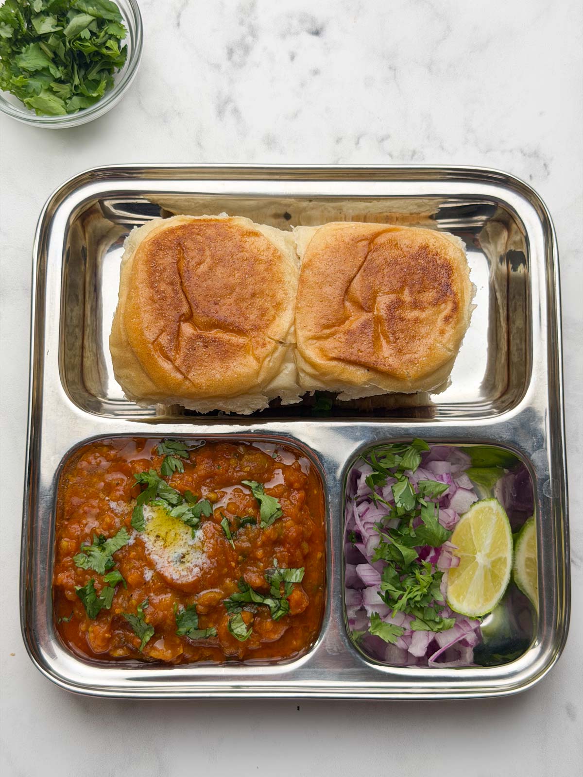 Pav and bhaji served in a bento steel plate with onion, coriander and lime wedge.