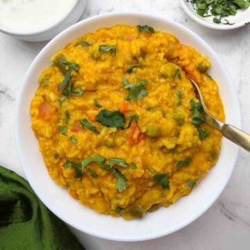 Masala vegetable khichdi served in a white bowl with a golden color spoon and yogurt and coriander leaves on the side.