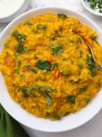 Masala vegetable khichdi served in a white bowl with a golden color spoon and yogurt and coriander leaves on the side.