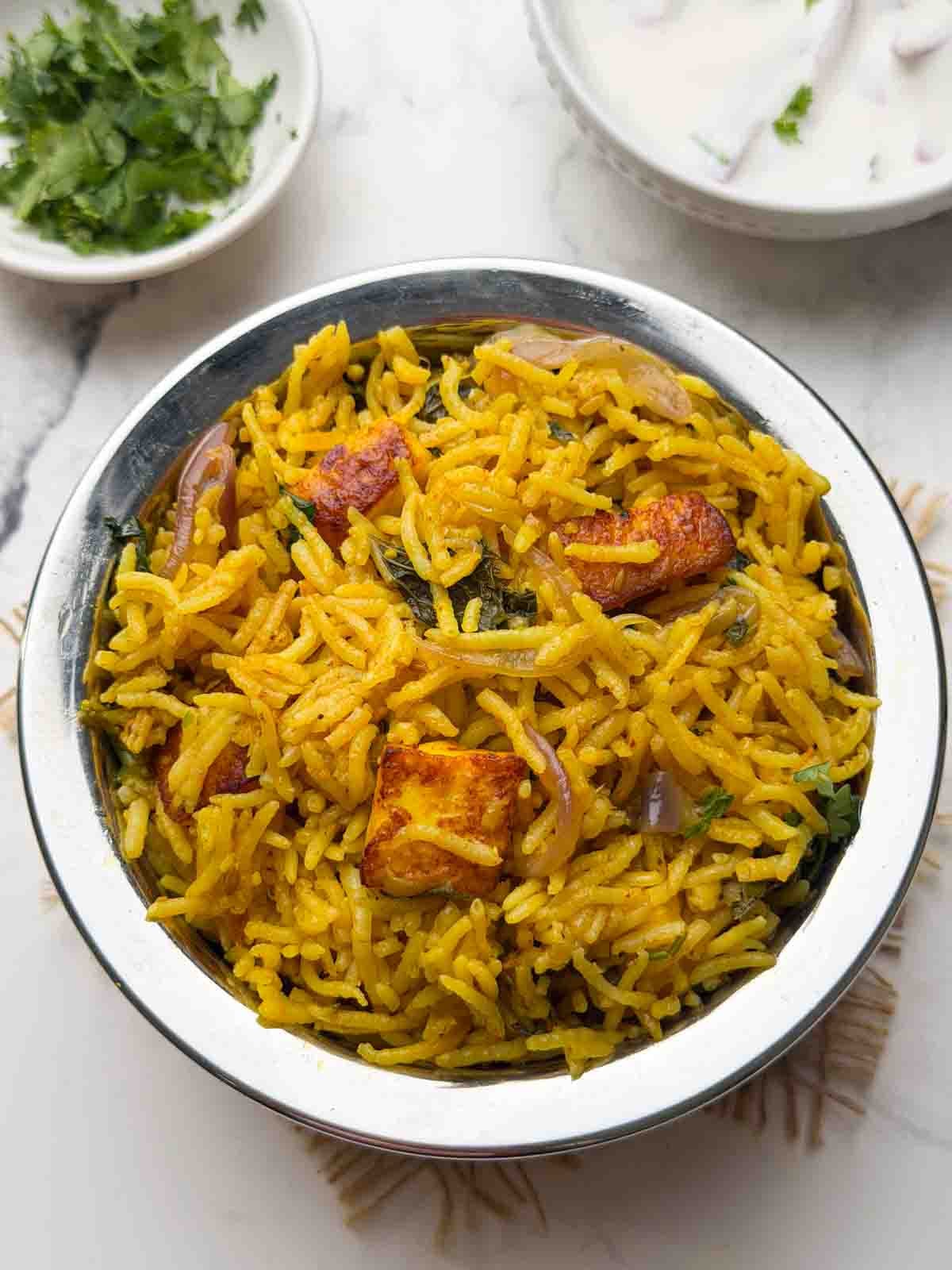 pressure cooker Paneer biryani in a steel serving pot with raita and coriander leaves on the side.