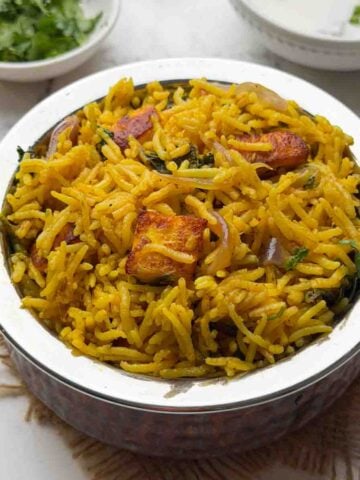 pressure cooker Paneer biryani in a steel serving pot with raita and coriander leaves on the side.