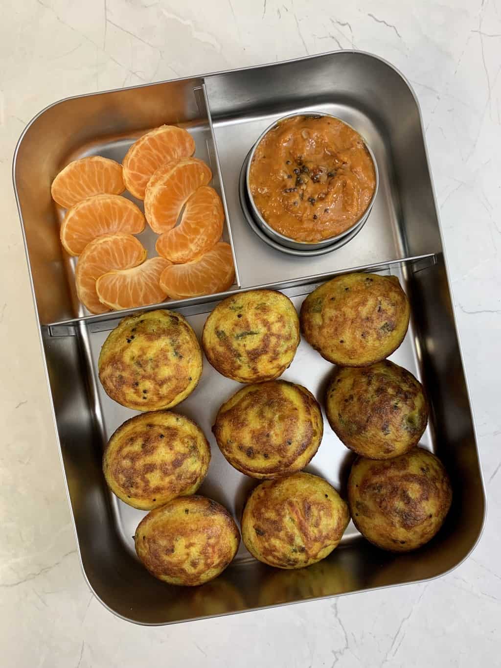 vegetable paniyaram, onion chutney and orange in bento steel lunch box.