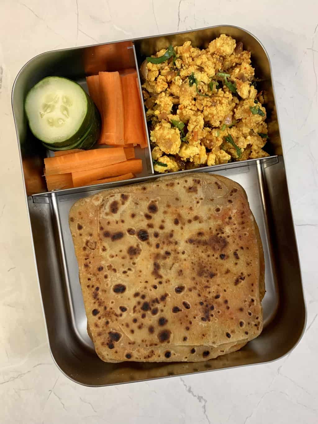 ajwain paratha, paneer bhurji, cucumber and carrot sticks served in a steel lunch box