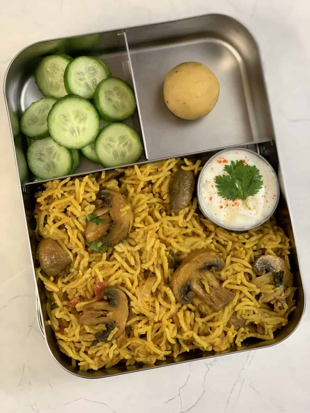 Mushroom Biryani, Yogurt, Besan Ladoo and Cucumber in steel lunch box.