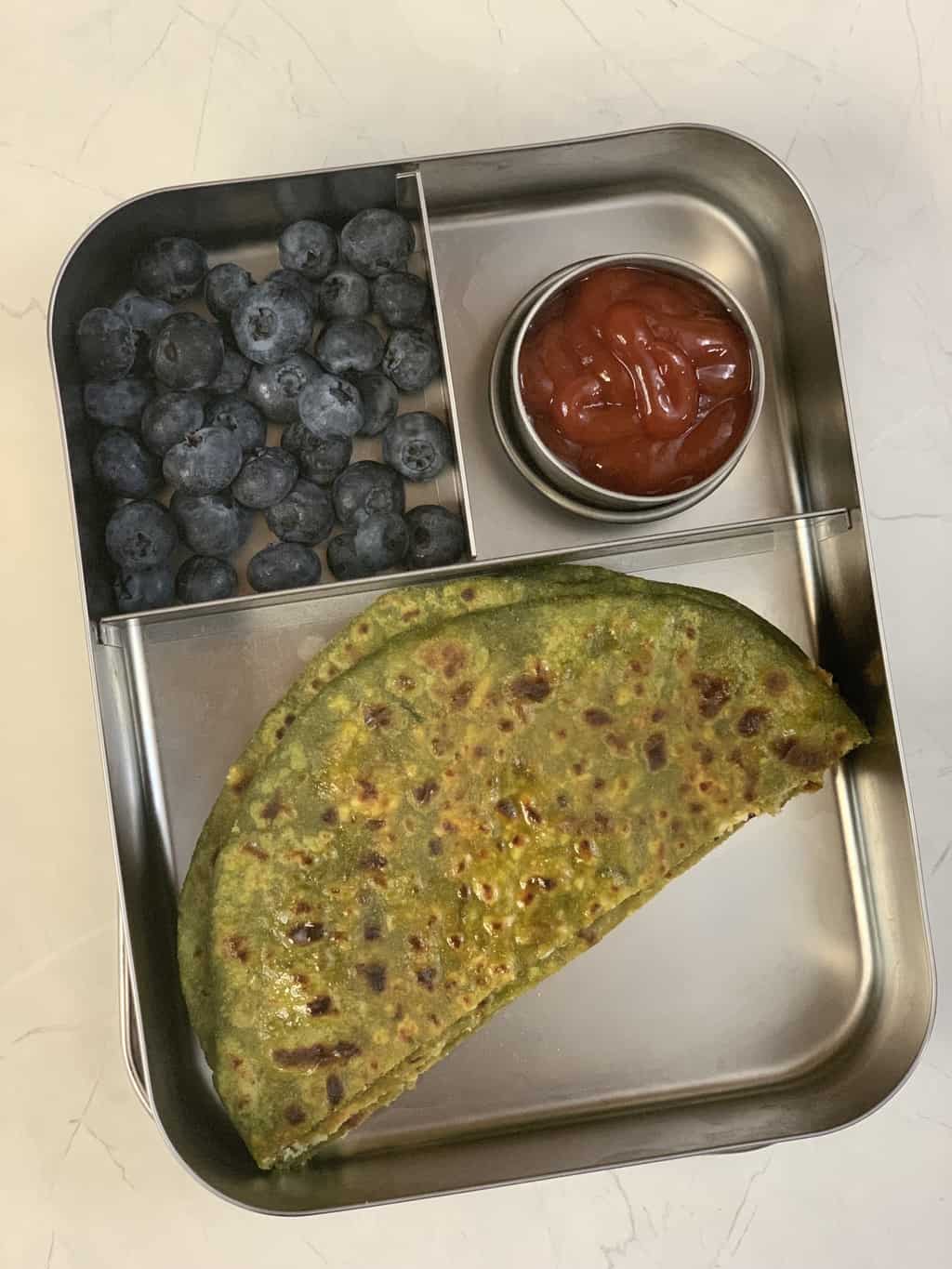 palak paneer paratha served in a kids lunch box with blueberries and ketchup