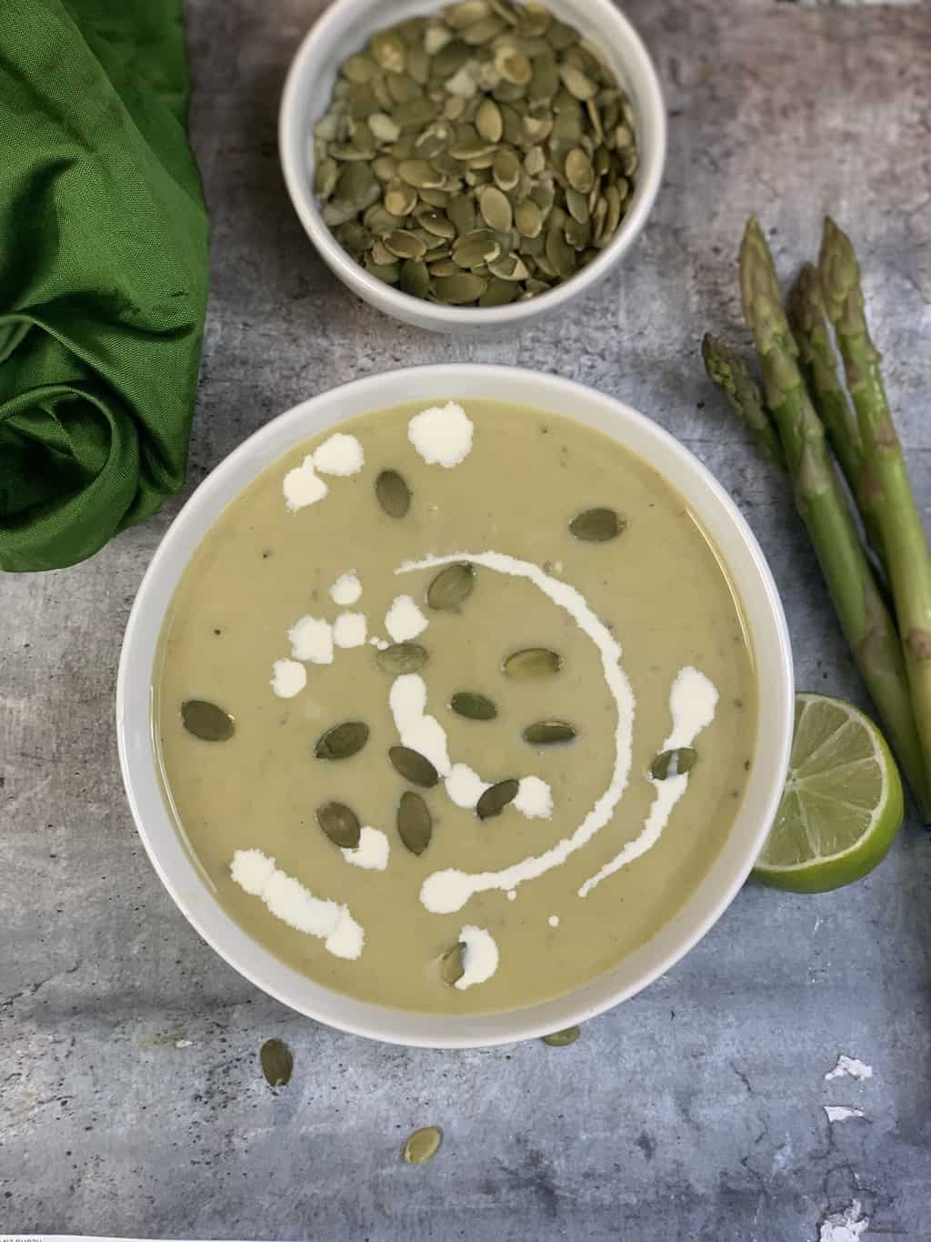 asparagus soup served in a bowl garnished with pumpkin seeds and cream with asparagus pumpkin seeds on the sides