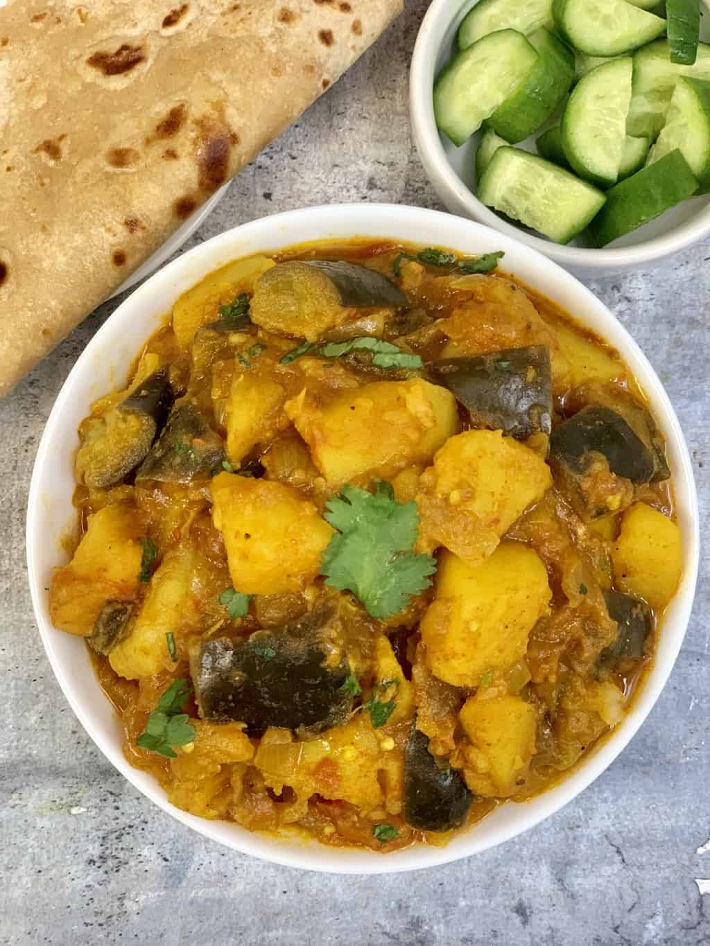 potato eggplant curry served in a bowl with chapati and chopped cucumber on the side