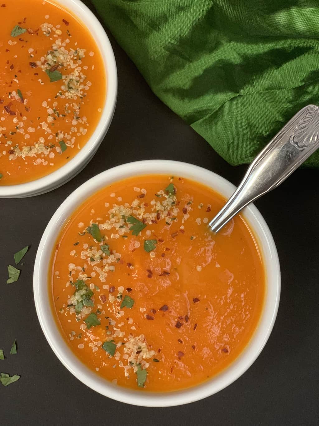 carrot ginger soup served in soup bowls with a spoon topped with hemp seeds chili flakes and cilantro