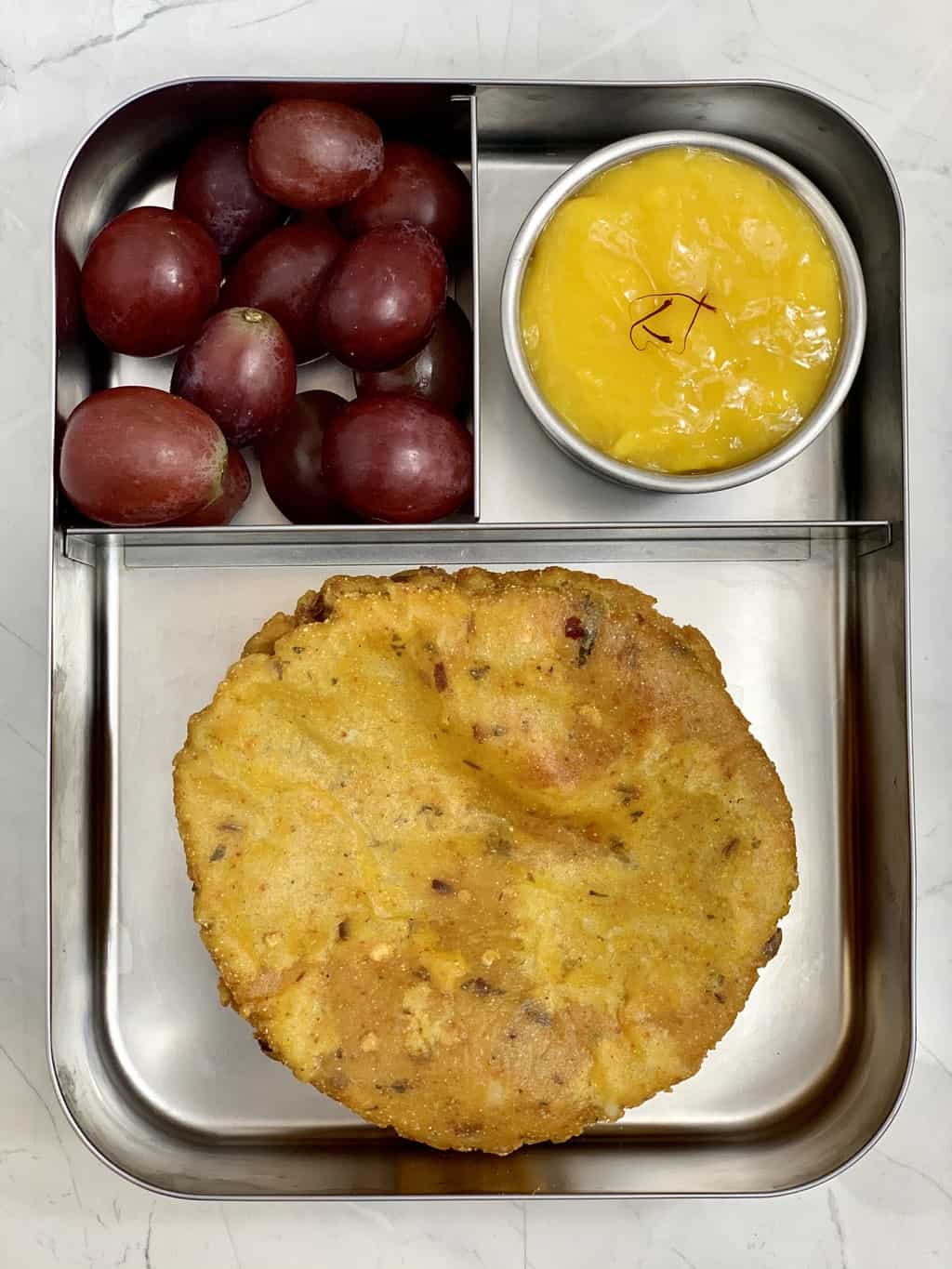 aloo masala poori with aamras and red grapes in steel lunch box.