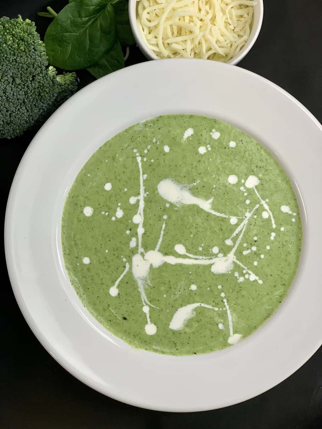 Broccoli Cheese Soup served in a soup bowl with cream on top and cheese and raw broccoli on the side