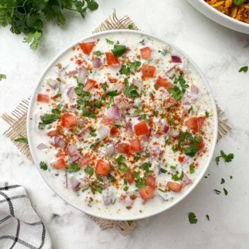 onion tomato raita served on a bowl garnished with roasted cumin powder, red chili powder with carrot rice and coriander bunch on the side