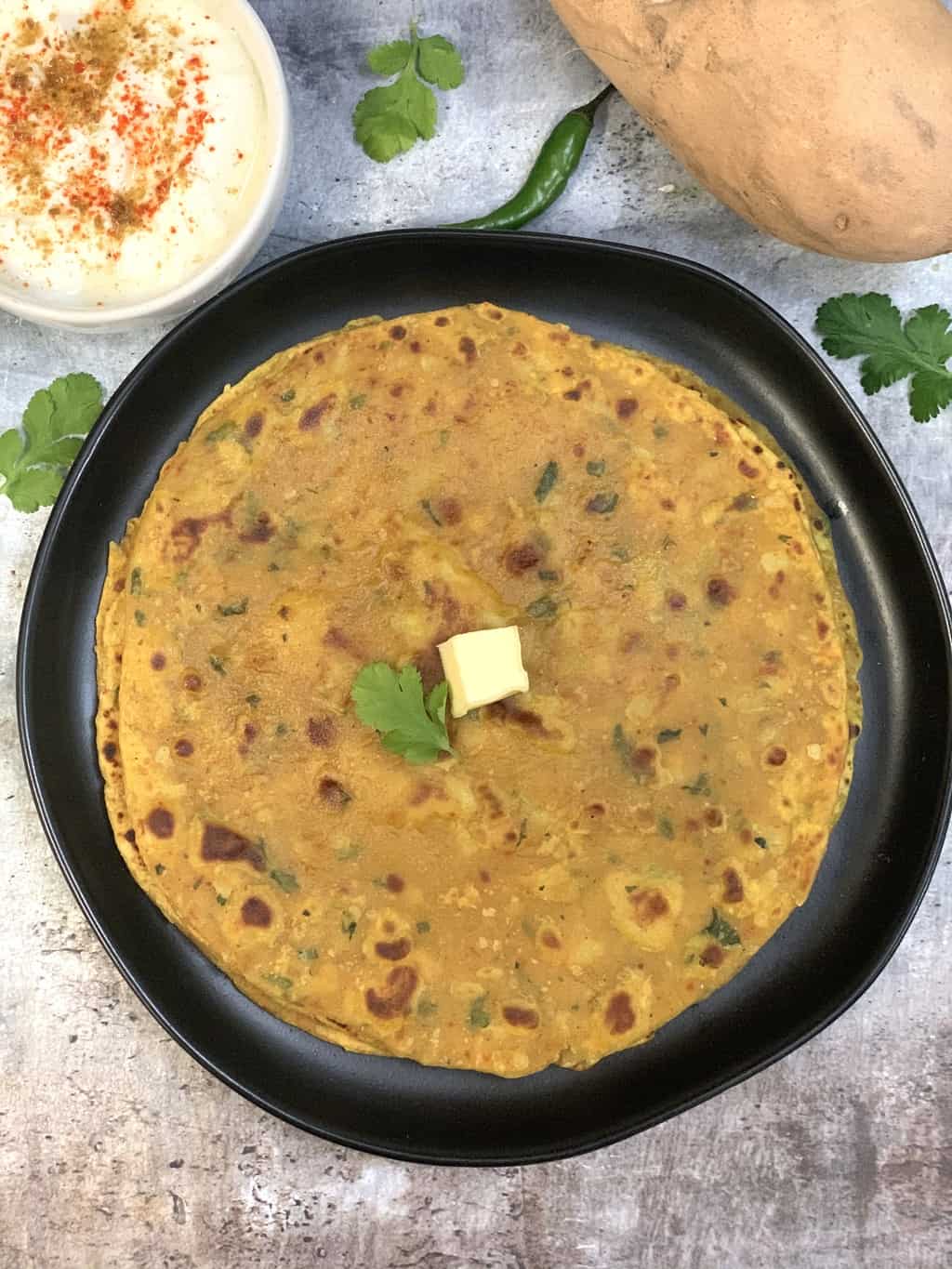 Sweet Potato Paratha stacked on a plate topped with butter and side of raita