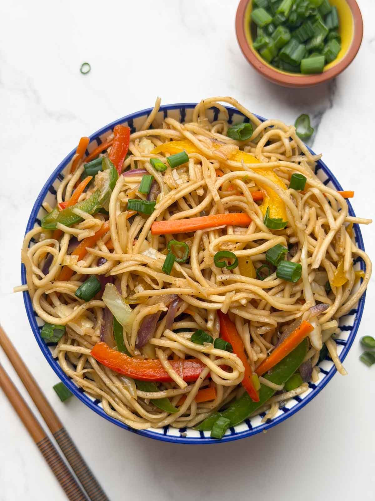 Vegetable hakka noodles served in a bowl, with side of chopped green spring onions, chings hakka noodles packet and chop sticks