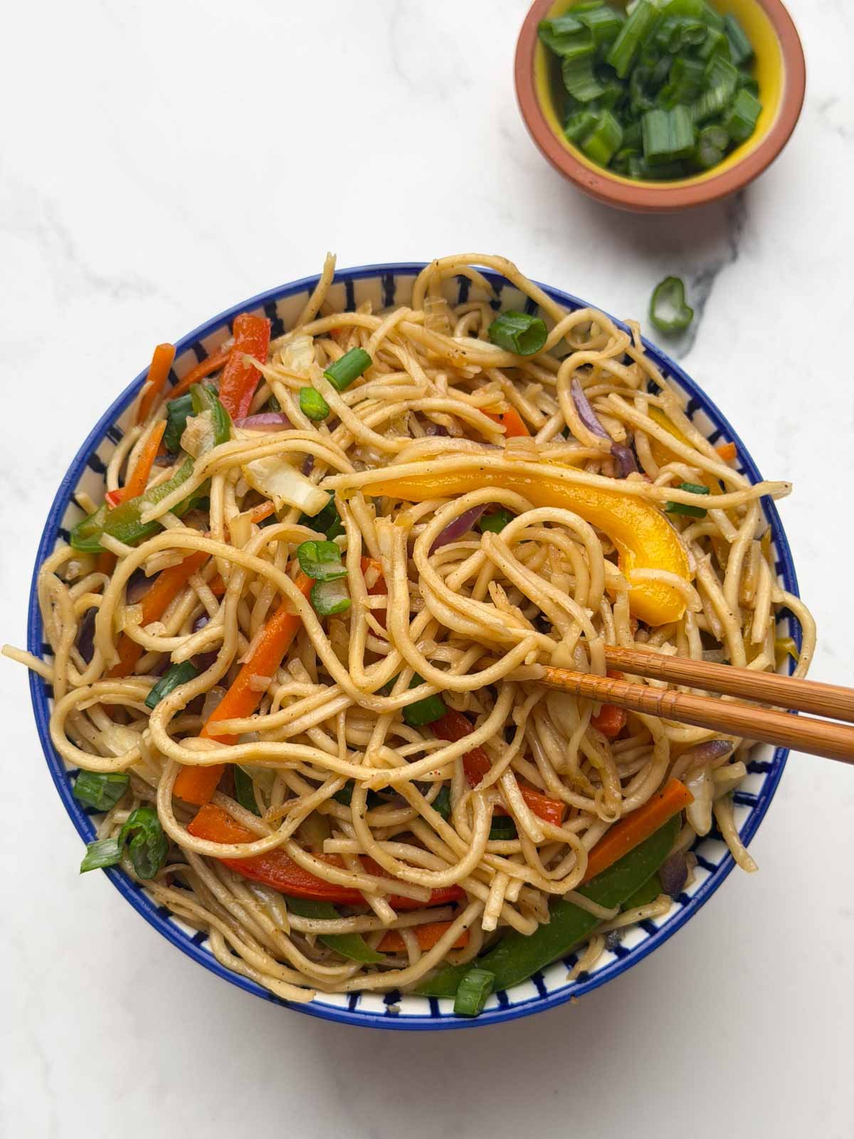 Vegetable hakka noodles served in a bowl, with with a pair of chopsticks lifting noodles, and chopped green spring onions on the side.
