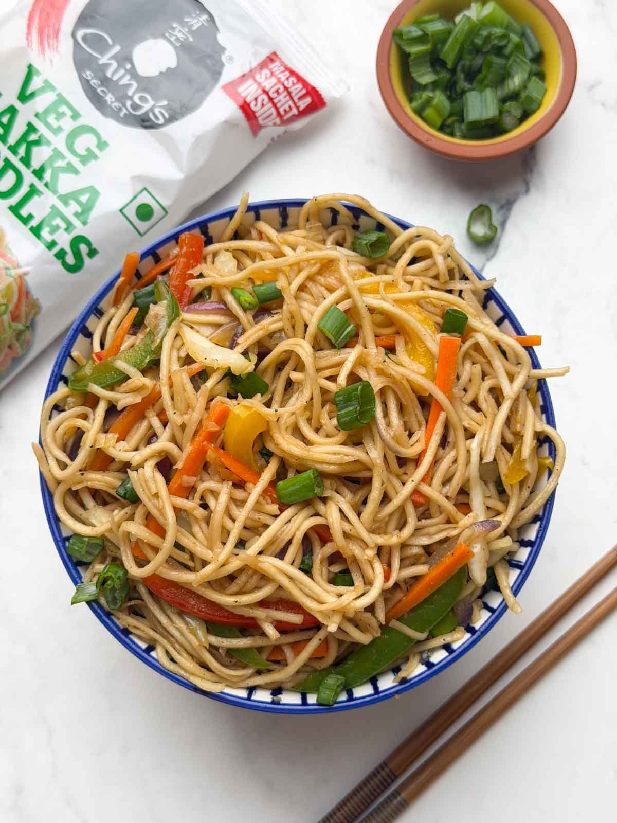 Vegetable hakka noodles served in a bowl, with side of chopped green spring onions, chings hakka noodles packet and pair of chopsticks placed on the side