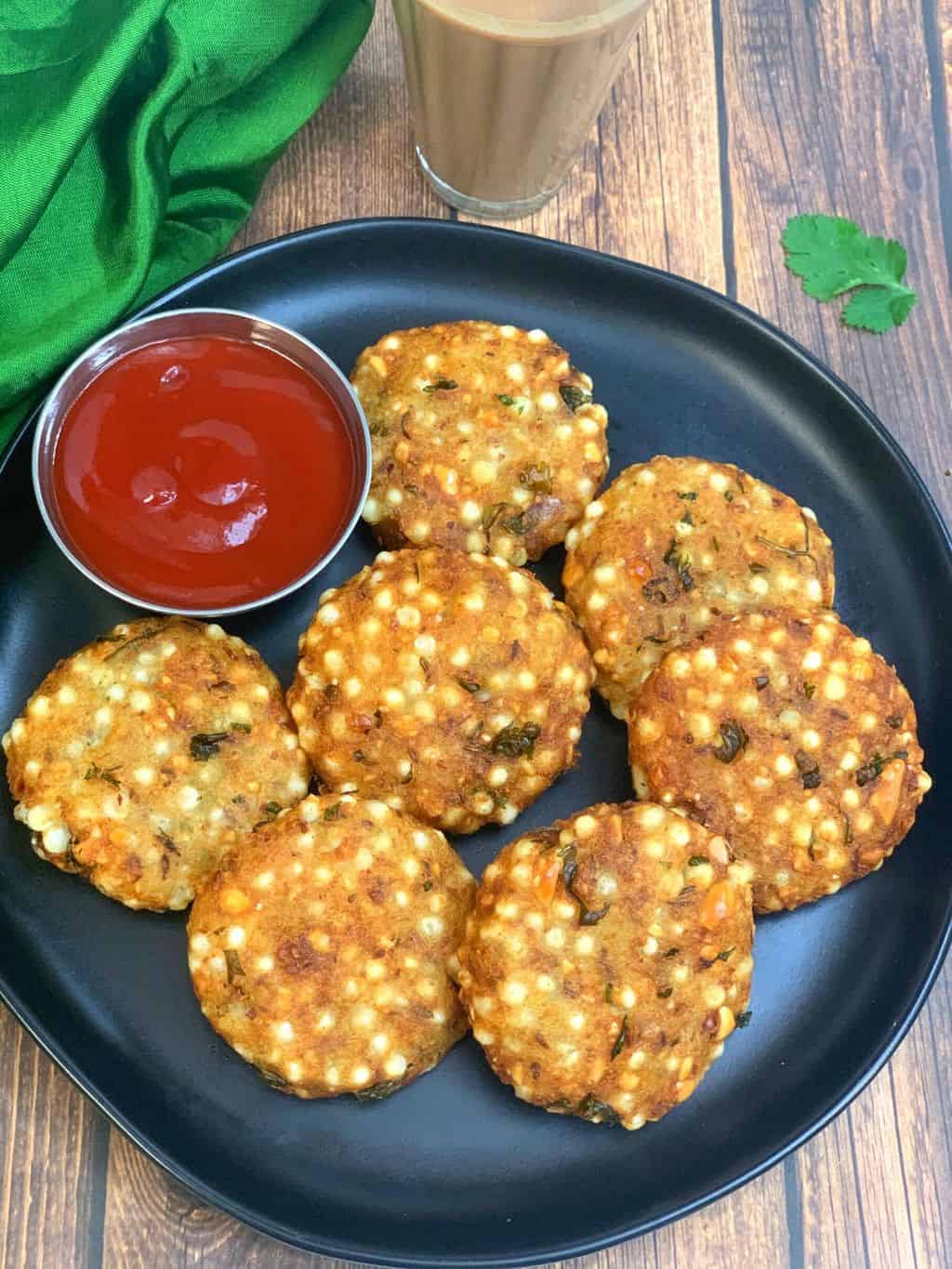 Sabudana vada served on a black plate with ketchup and tea on the side