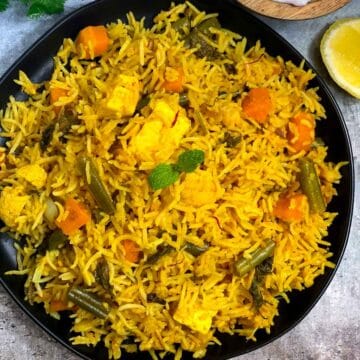 pressure cooker vegetable biryani served in a black plate with rait and lemon wedges on the side