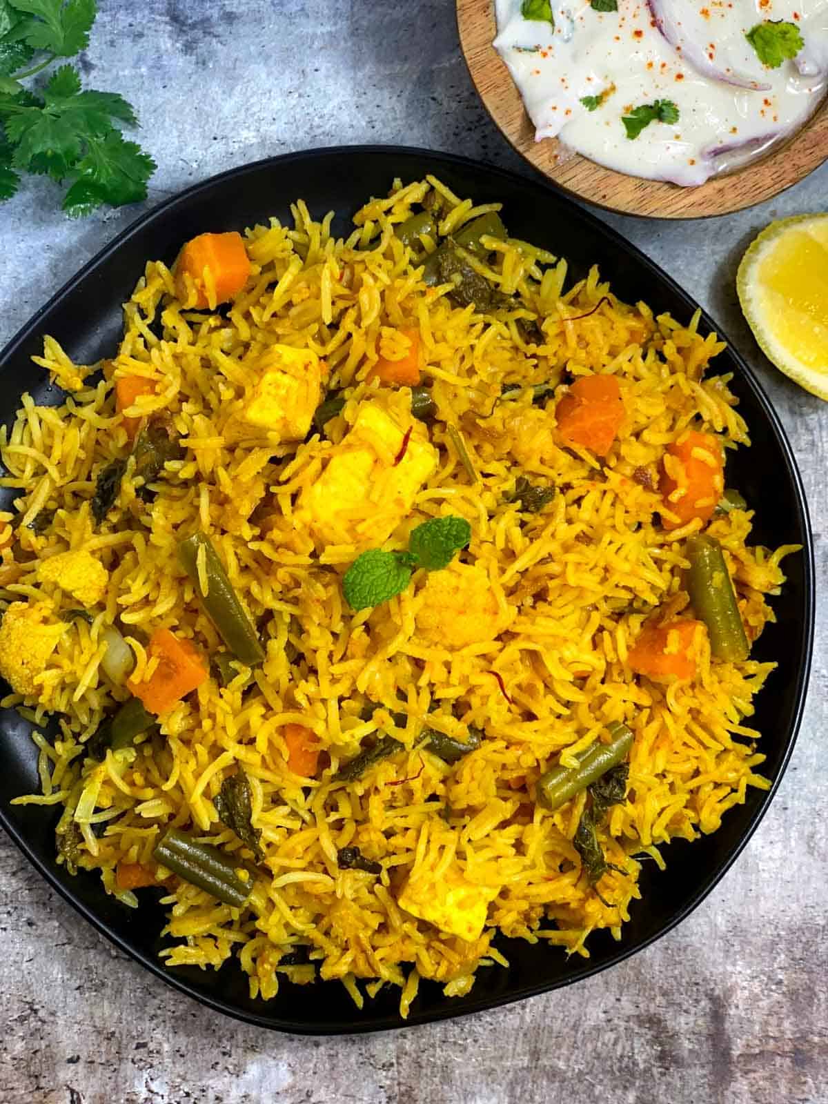 instant pot vegetable biryani served in a black plate with rait and lemon wedges on the side