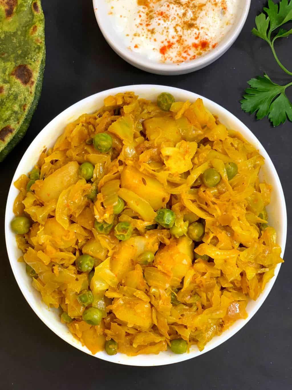 cabbage potato peas curry served in a bowl with paratha and raita on the side