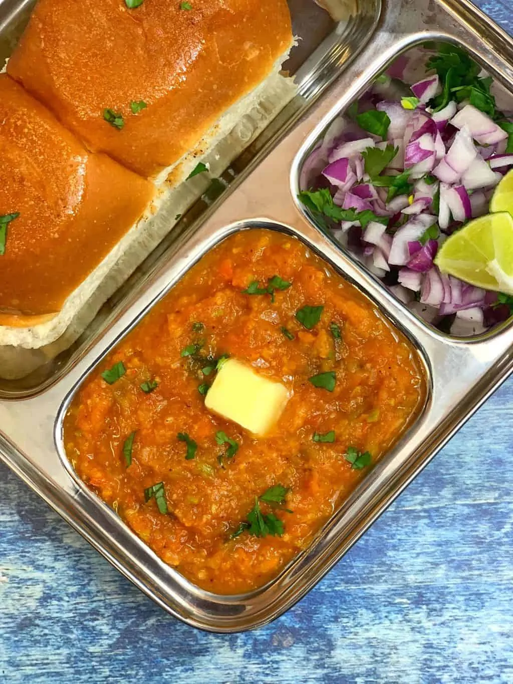pav bhaji served in a steel plate topped with butter and pav and onion lemon on the side