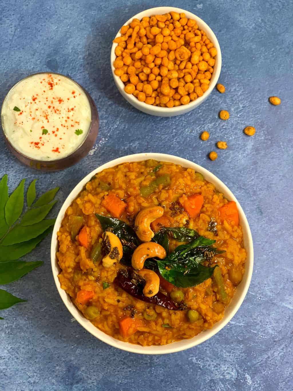 bisi bele bath served in a bowl with tempering on the top and raita and boondi on the side