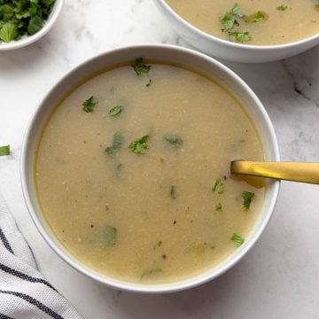 Lauki Moong Dal Soup served in two white bowl with a golden spoon and coriander leaves in a small bowl for garnish.