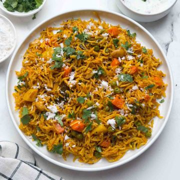 Masale bhaat served in a white plate accompanied by raita in a small bowl and coriander leaves on the side.
