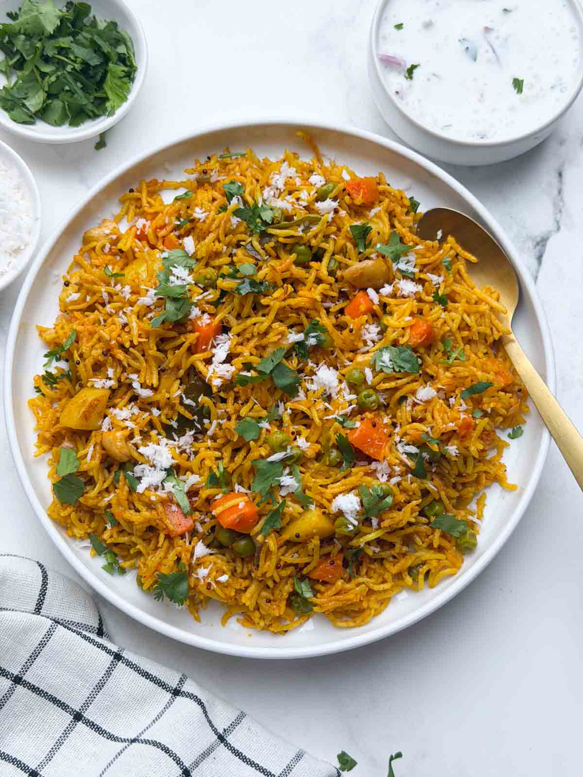 Masale bhaat served in a white plate with a golden spoon on it accompanied by raita in a small bowl and coriander leaves on the side.