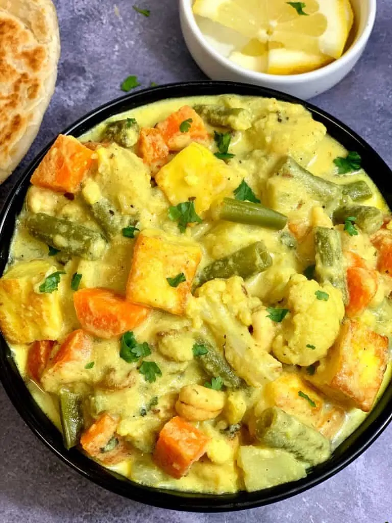 Navratan Korma served in a black bowl garnished with cilantro with paratha and lemon wedges on the side