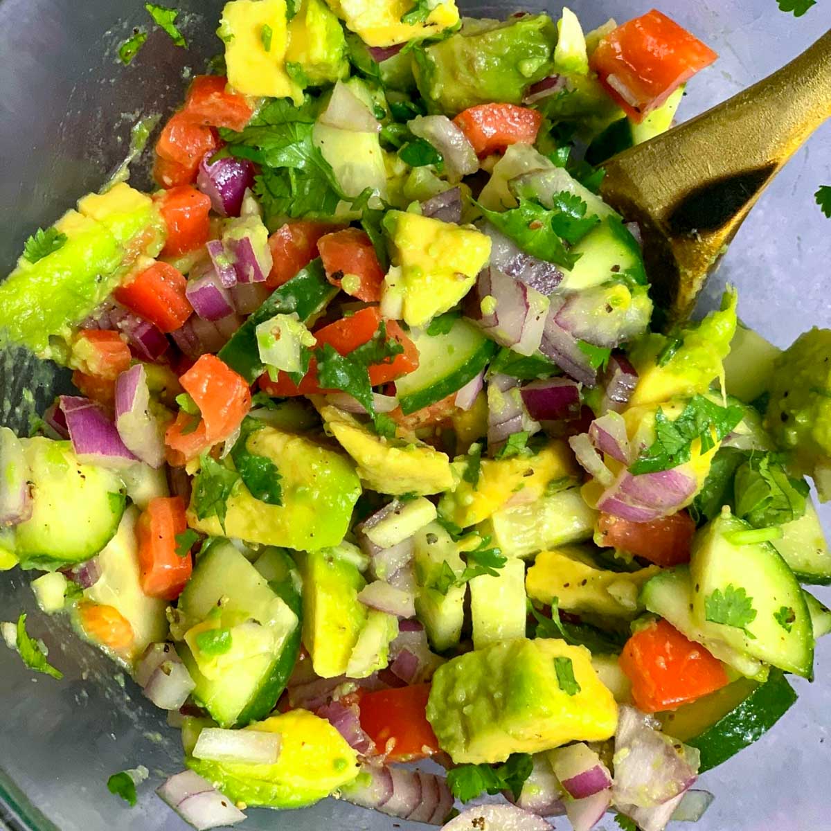 avocado cucumber tomato salad served in a glass bowl with wooden spoon