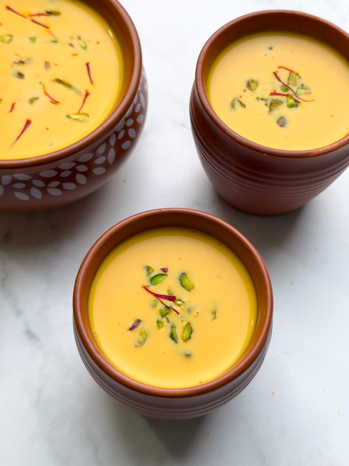 Carrot kheer in 2 mugs and 1 large pot garnished with saffron and pistachios.