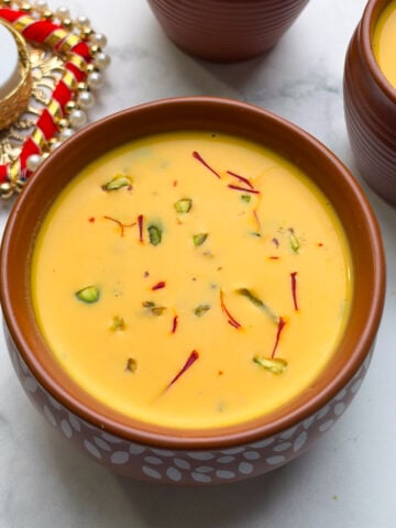 Carrot almond kheer served in a pot, topped with chopped pistachios and saffron, and two more mugs of kheer on the side with decorative candle.