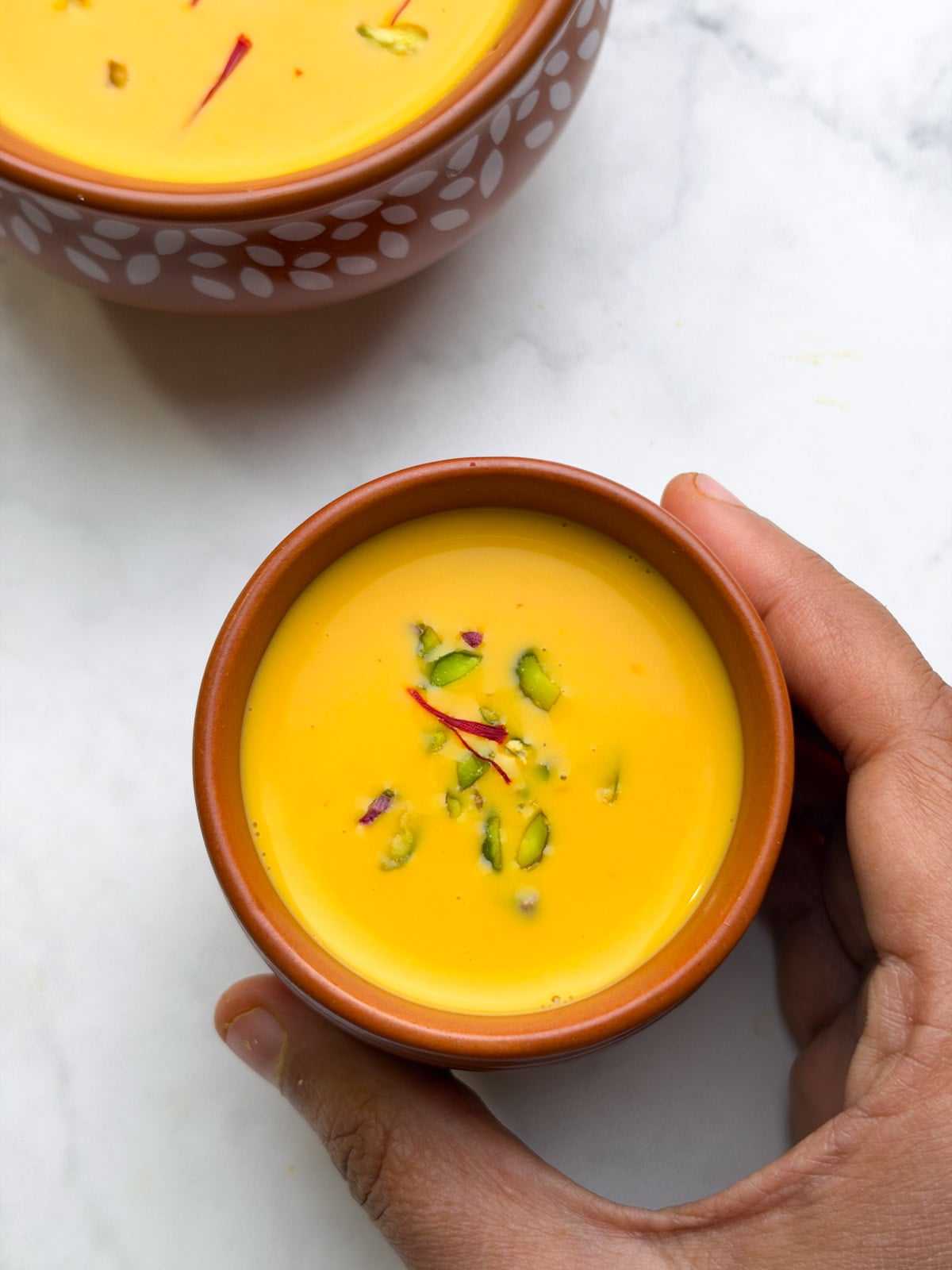 Carrot almond kheer served in a mug, topped with chopped pistachios and saffron, held by hand and another pot of kheer on the side.