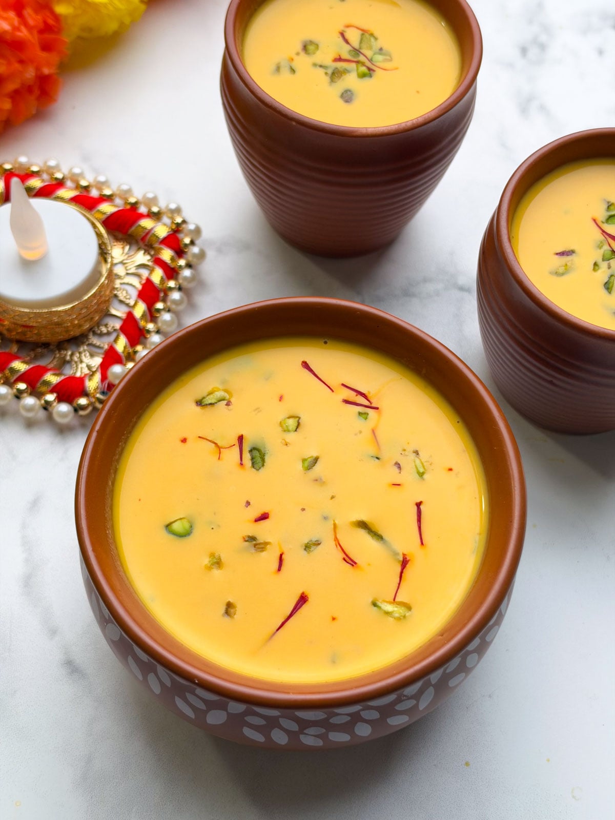 Carrot almond kheer served in a pot, topped with chopped pistachios and saffron, and two more mugs of kheer on the side with decorative candle