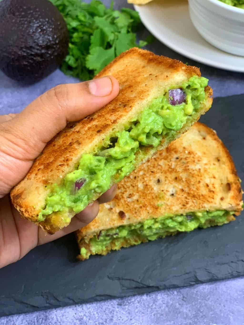 grilled guacamole sandwich served on a black slate with one of the slice held in a hand 