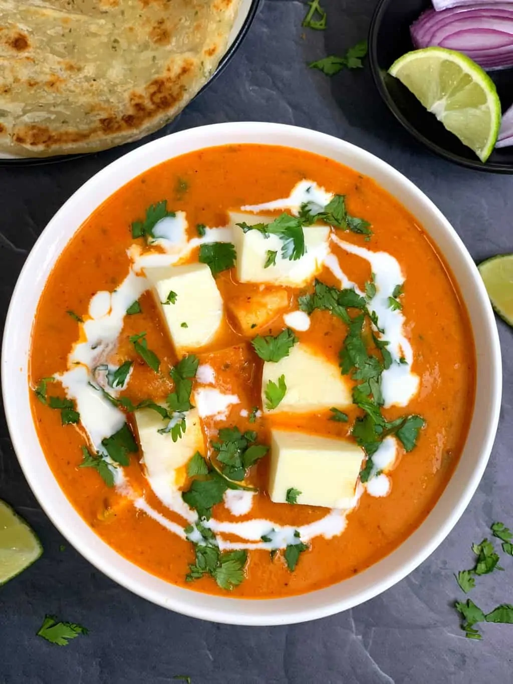 Restaurant Style Paneer Butter Masala in a white bowl with cream and coriander leaves for garnish with paratha on side with lemon wedges.
