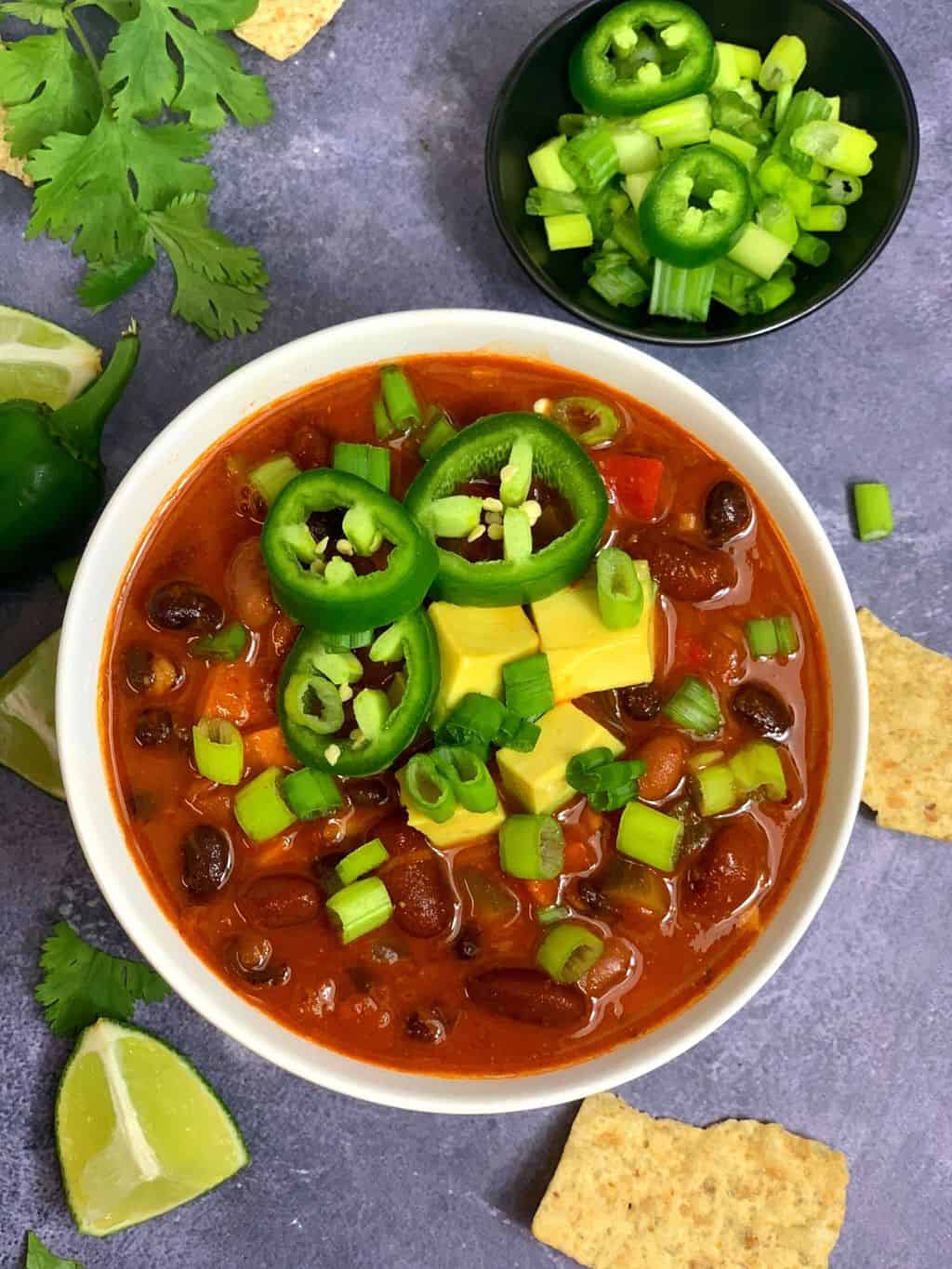 Three bean vegetarian chili cooked in instant pot served in a white bowl with dpring onions ,lemon wedges ,tortila chips on side