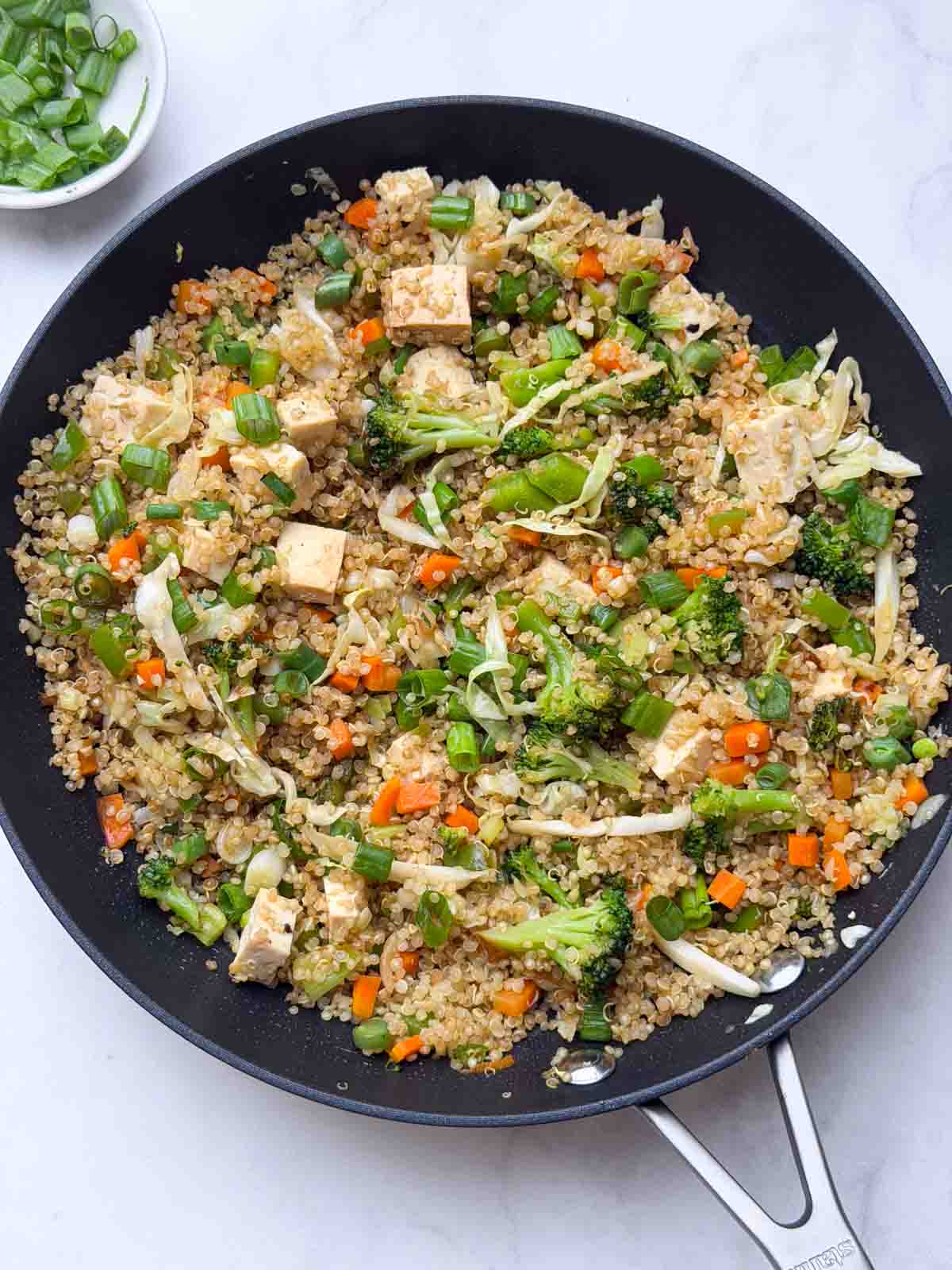 Quinoa vegetable fried rice in a black pan with green spring onions on the side.