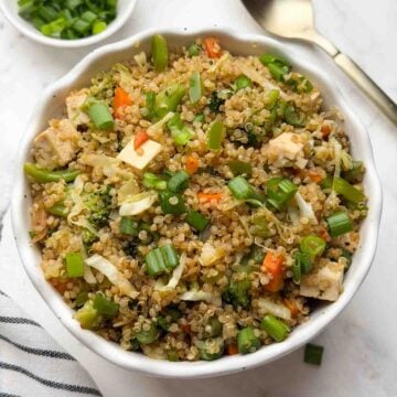 Quinoa fried rice served in a white bowl with spring onions and spoon on the side.