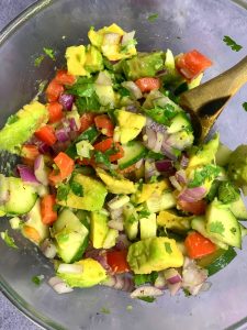 avocado cucumber tomato salad in a glass bowl with a wooden spoon